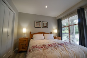 A bedroom with a bed, two bedside tables, a nightstand, a window with blinds, and two framed pictures on the wall. - Stoney Creek Northstar 8 (Whistler)