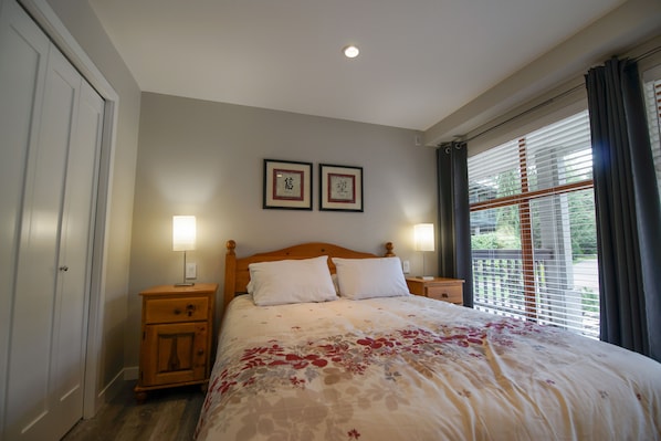 A bedroom with a bed, two bedside tables, a nightstand, a window with blinds, and two framed pictures on the wall. - Stoney Creek Northstar 8 (Whistler)