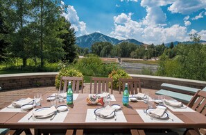 An outdoor dining area with tables set for four, each with plates, cutlery, and glasses. There are two bottles of Pellegrino water on the tables. The setting overlooks a scenic landscape with trees, a river, and mountains in the distance. - Captivating Historic Cottage with Fireplace (Sun Valley)