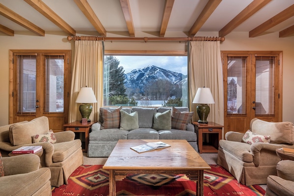 Ein gemütliches Wohnzimmer mit einer Sofagarnitur, einem Couchtisch und Lampen. Ein Fenster bietet Blick auf schneebedeckte Berge.