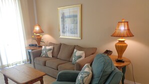 A living room with a sofa, armchair, and a small table. There are two lamps on side tables, and a framed picture on the wall. - The Inn at Dauphin Island 102 Walk right out to Pools and Beach! (Dauphin Island)