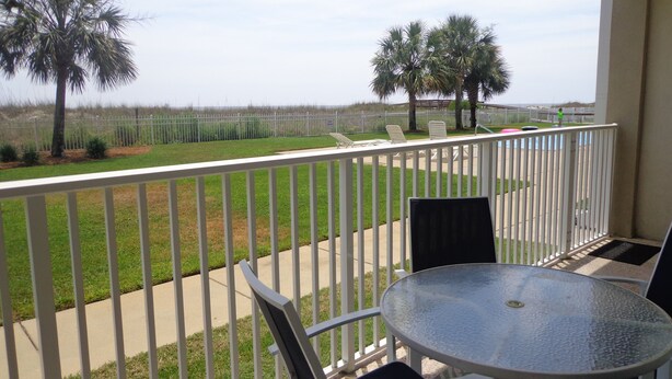 A balcony with a round table and chairs, overlooking a grassy area with palm trees and a fence. - The Inn at Dauphin Island 102 Walk right out to Pools and Beach! (Dauphin Island)