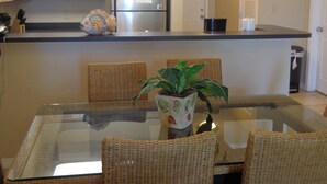 A dining area with a glass table surrounded by wicker chairs, a chandelier, and a refrigerator in the background. - The Inn at Dauphin Island 102 Walk right out to Pools and Beach! (Dauphin Island)