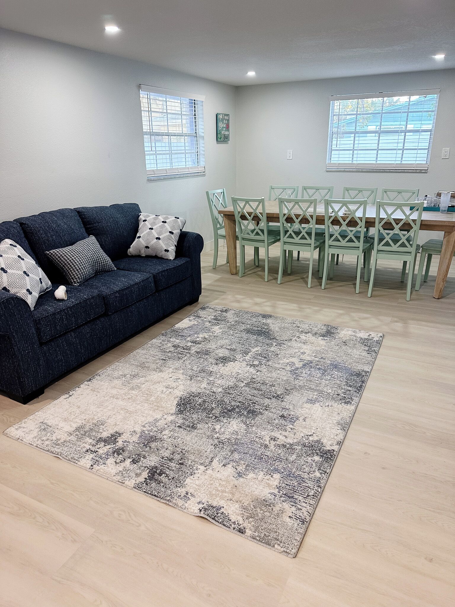 A living room with a dark blue sofa, a patterned rug, a dining table with green chairs, and two windows with blinds.