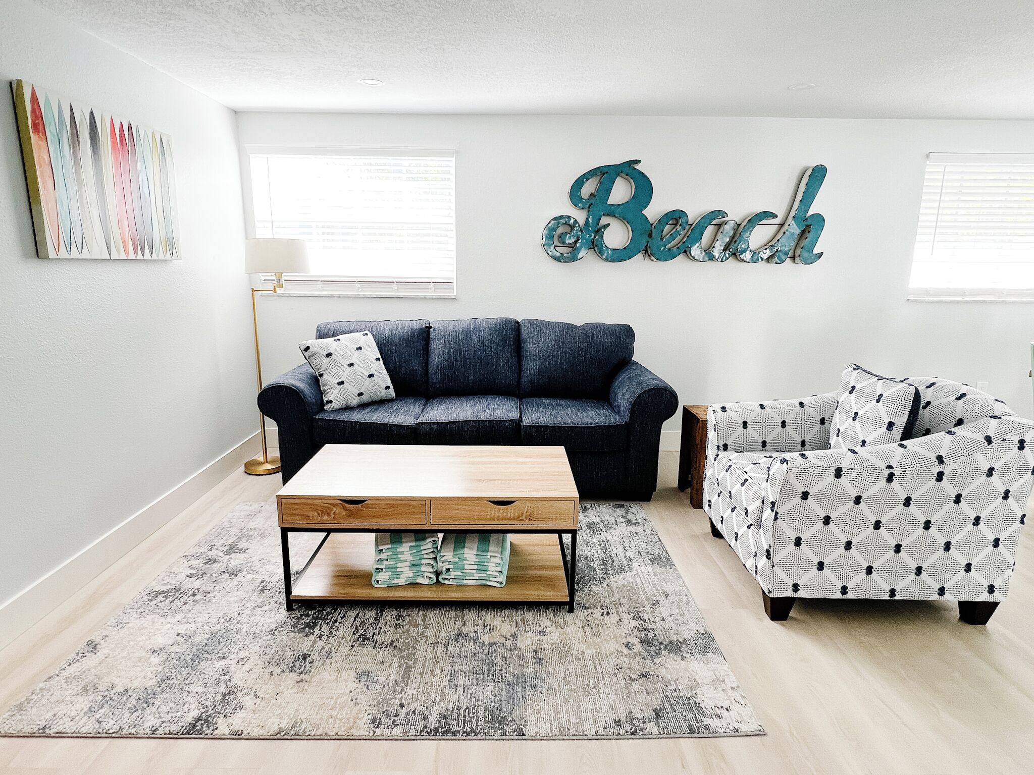 Ein Wohnzimmer mit einem dunkelblauen Sofa, einem gemusterten Sessel und einem Holztisch. Eine große Wanddekoration zeigt das Wort 'Beach'.