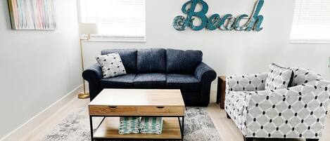 Ein Wohnzimmer mit einem dunkelblauen Sofa, einem gemusterten Sessel und einem Holztisch. Eine große Wanddekoration zeigt das Wort 'Beach'.