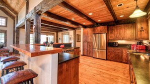 A rustic kitchen with wooden beams, a central island with stools, stainless steel appliances, and a large window. - Mountain View | Hot Tub | Game Room (Big Sky)