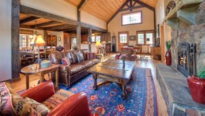 A spacious living room with a high ceiling, wooden beams, a stone fireplace, and a large window. - Mountain View | Hot Tub | Game Room (Big Sky)
