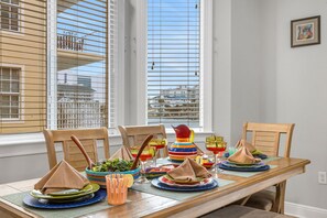Une table à manger dressée avec de la vaisselle et des serviettes aux couleurs vives, des chaises en bois et une fenêtre avec des stores.