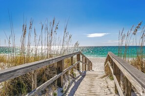 A wooden boardwalk leading to a beach with clear blue water and tall grasses.