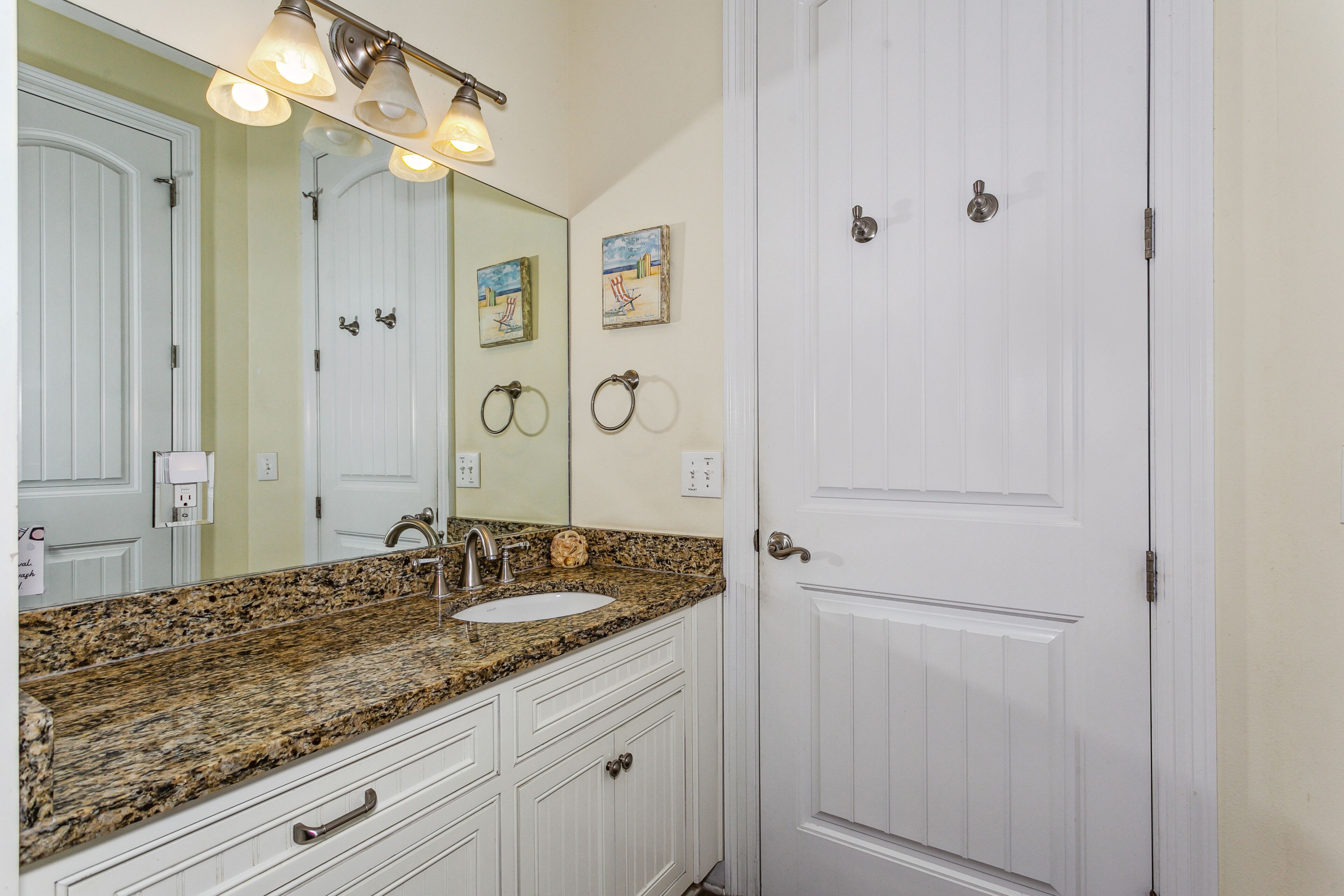 A bathroom with a granite countertop sink, white cabinetry, a large mirror, and a white door.
