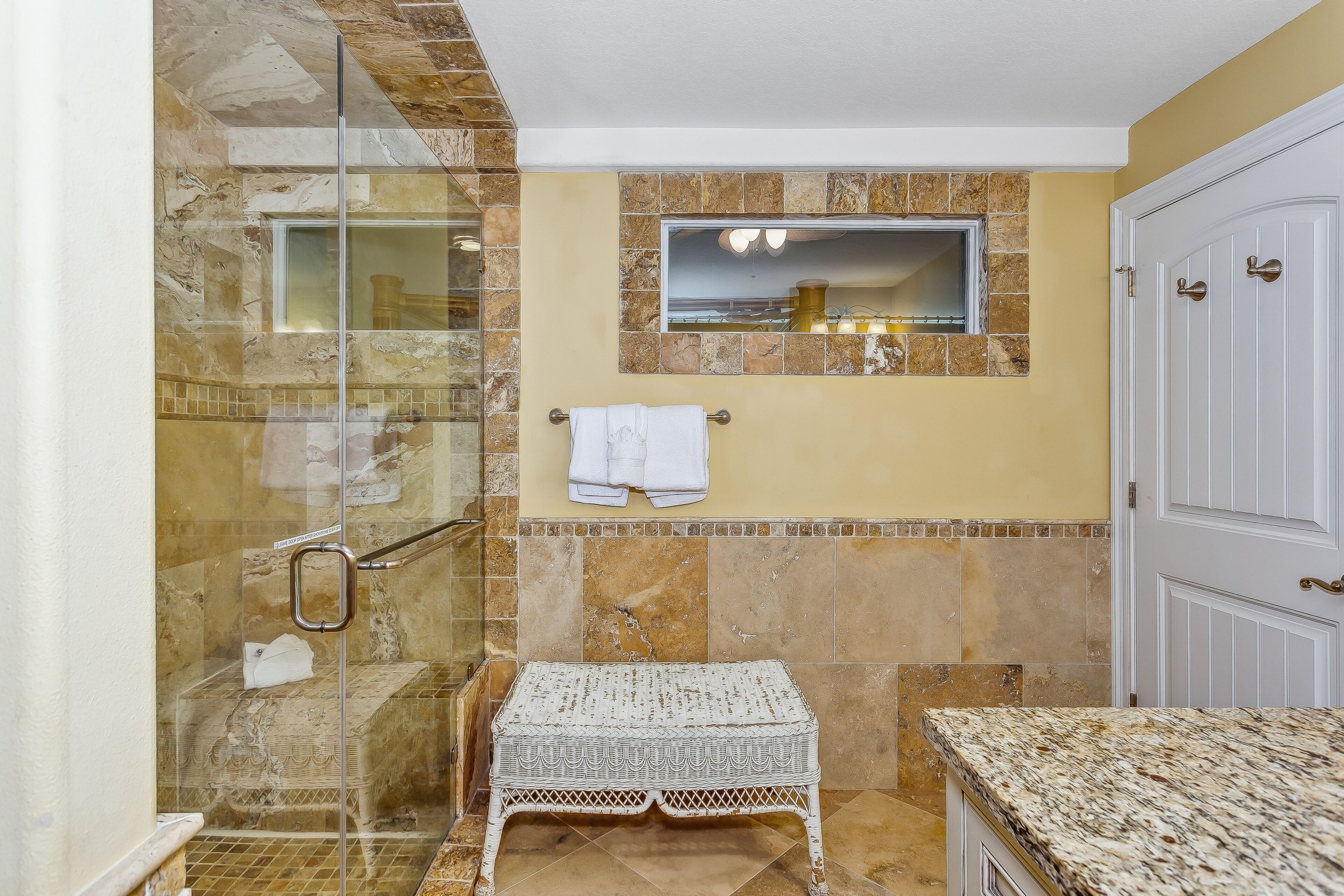 A bathroom with a glass shower enclosure, a white bench, a towel rack, and a door.