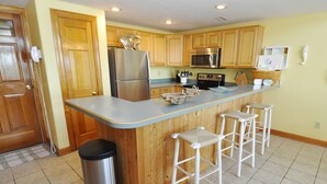 A kitchen with wooden cabinets, a stainless steel refrigerator, a microwave, and a sink. There are three white bar stools and a trash can. - Almost Heaven: Colorful sunsets and views of Portsmouth Island. (Ocracoke)