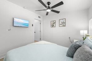 A bedroom with a bed, a ceiling fan, a TV, and two framed pictures on the wall. - Sandy Shores • Steps to Beach & Frenchy's • Pet Friendly • Patio ➠ 20 Kendall 1 (Clearwater)