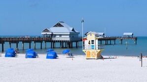 Ein Strand mit einem Rettungsturm, blauen Sonnenschirmen und einem Steg mit Gebäuden.