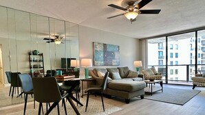 Ein modernes Wohnzimmer mit Glastrennwand, Deckenventilator, Essbereich mit Stühlen, einer Couch und einem großen Fenster mit Stadtblick.
