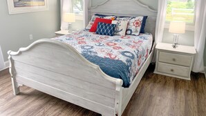 A bedroom with a white bed, nautical-themed bedding, a small bedside table with a lamp, and a wall-mounted picture. - Pelican's Perch: Centrally located Dog friendly condo with pool. (Ocracoke)