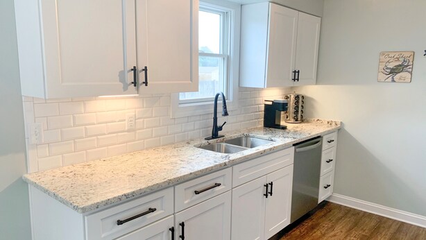 A kitchen with white cabinets, a granite countertop, a sink, a coffee maker, and a window. - Pelican's Perch: Centrally located Dog friendly condo with pool. (Ocracoke)
