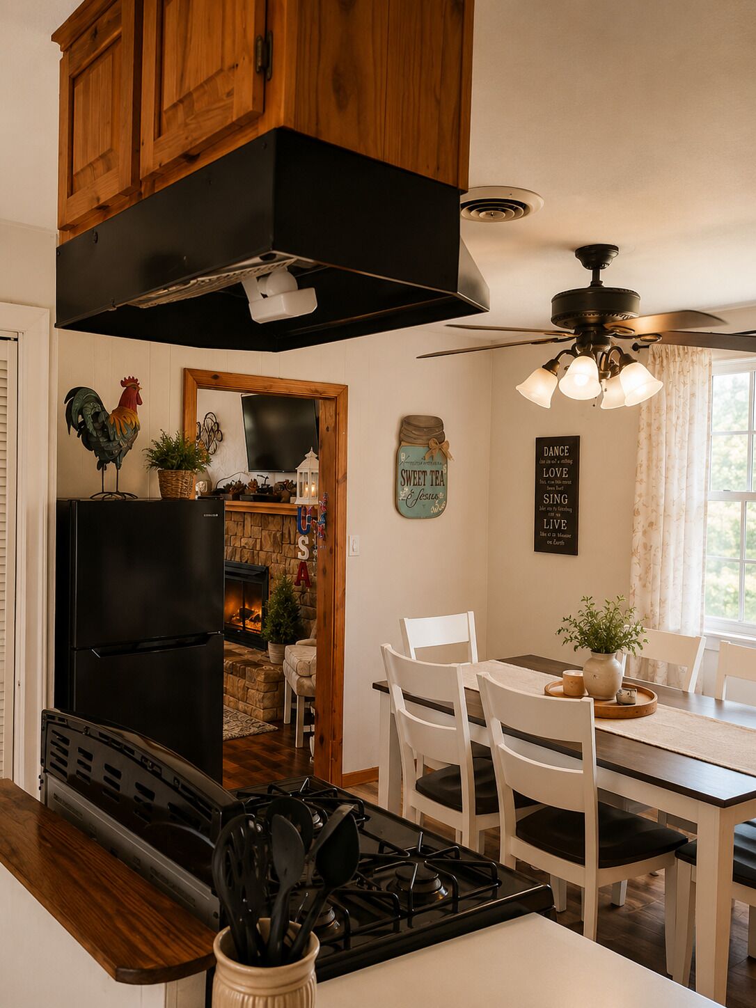 A kitchen with a black refrigerator, wooden cabinets, a stone fireplace, a dining table with white chairs, and a ceiling fan.