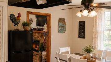 A kitchen with a black refrigerator, wooden cabinets, a stone fireplace, a dining table with white chairs, and a ceiling fan.
