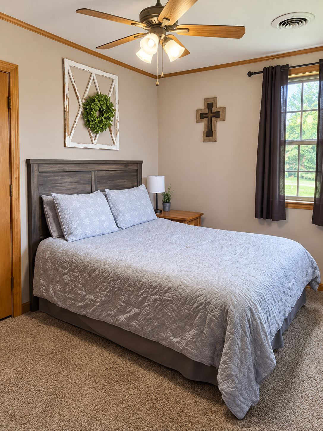 A bedroom with a bed, a ceiling fan, a cross on the wall, and a window with curtains.