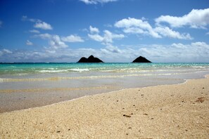 Een strand met helder turquoise water en twee donkere rotsformaties in de verte.