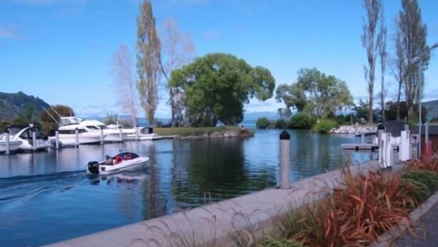 Una marina con barcos amarrados, un camino a lo largo del agua y árboles al fondo.