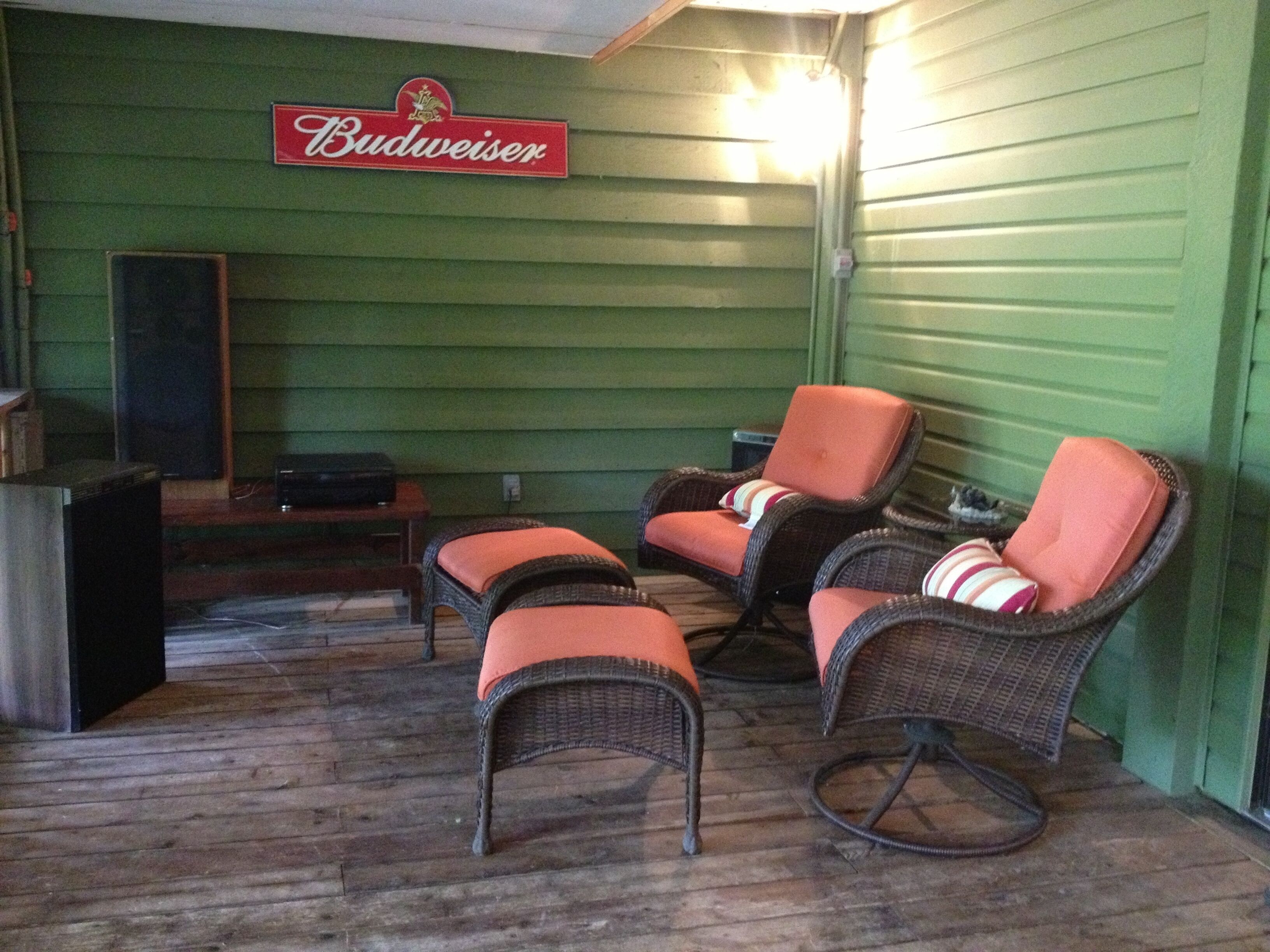 A room with green walls, a sign reading "Budweiser," wicker chairs with orange cushions, a wooden table, and a speaker.