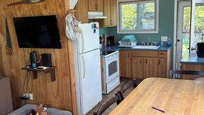 A compact kitchen with wooden cabinets, a white refrigerator, a stove, and a sink. There is a dining table with chairs and a TV mounted on the wall. - Summer family cabin on Lake Ontario, Pulaski
NEWLY UPDATED!! (Pulaski)