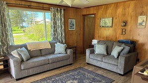 A living room with two grey sofas, a dark patterned rug, wooden walls, and a view of greenery outside. - Summer family cabin on Lake Ontario, Pulaski
NEWLY UPDATED!! (Pulaski)