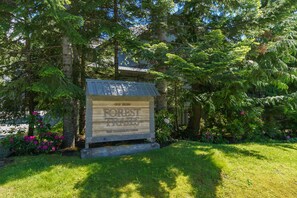 A wooden sign for 'Forest Trails' with a metal roof, surrounded by lush greenery and flowering plants. - Forest Trails - Easy Ski Access, Private Garage, Free Village Shuttle (Whistler)