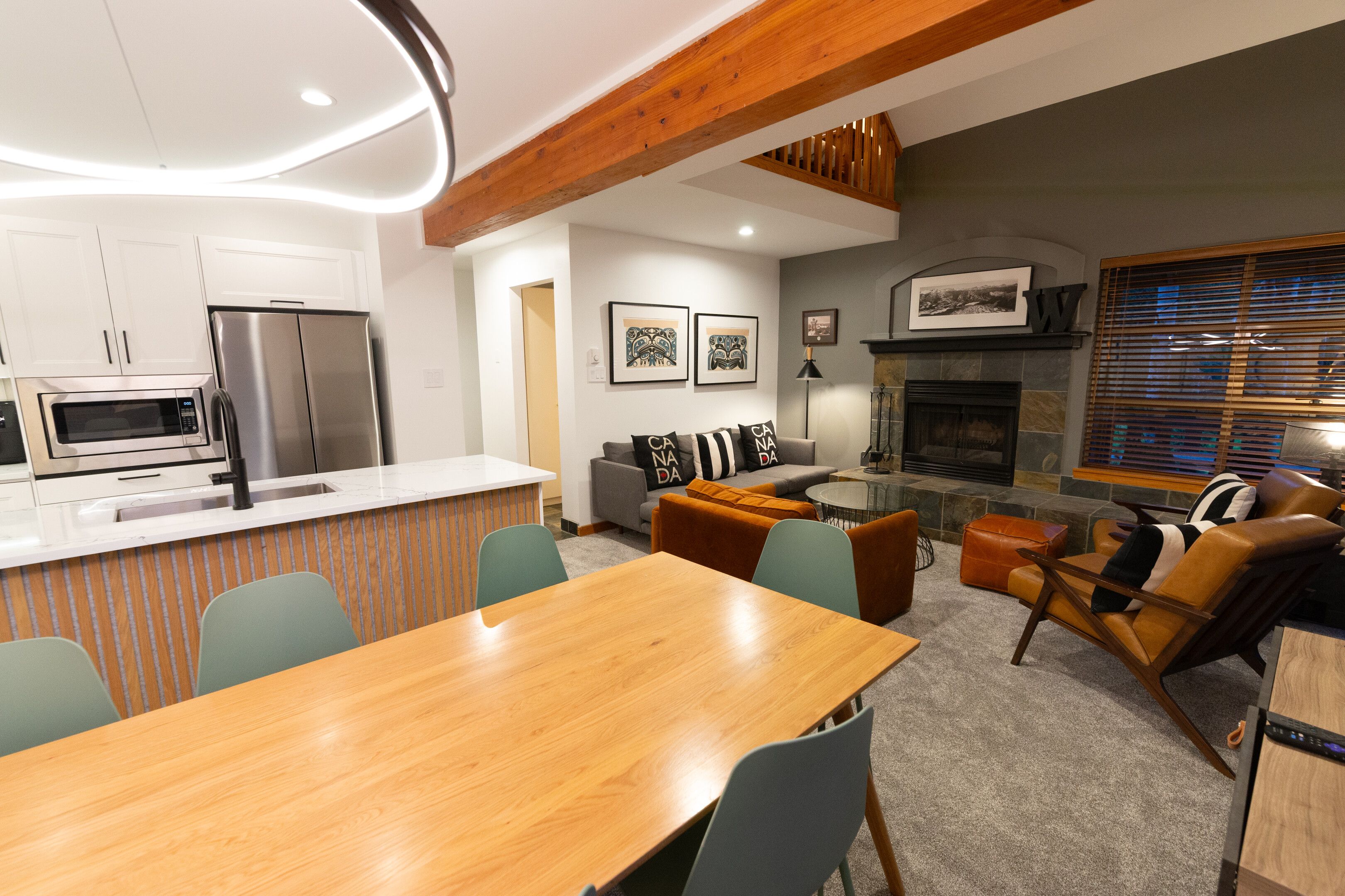 A modern kitchen with stainless steel appliances, a dining area with a wooden table and chairs, a living room with a fireplace, and a view of the outdoors through large windows.