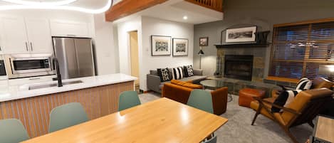 A modern kitchen with stainless steel appliances, a dining area with a wooden table and chairs, a living room with a fireplace, and a view of the outdoors through large windows.