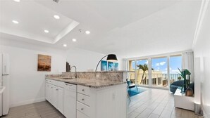 Cozinha moderna com armários brancos, bancada de granito e uma ilha grande. Há uma área de jantar com um sofá azul e vista para o exterior.