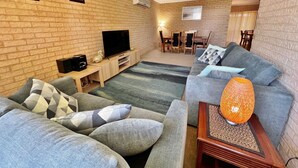 Ein Wohnzimmer mit Backsteinwand, einem Fernseher, einer Couch und einem Essbereich im Hintergrund.