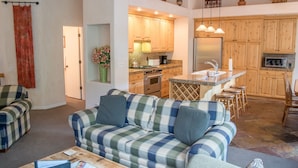 A cozy living room with a plaid sofa, a coffee table, and a kitchen area with wooden cabinets and a stainless steel refrigerator. - River Ridge Townhomes - Sleek Family Foxhole Retreat with Private Hot Tub (Ketchum)