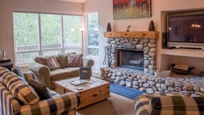 A cozy living room with a stone fireplace, a large window, and a television. - River Ridge Townhomes - Sleek Family Foxhole Retreat with Private Hot Tub (Ketchum)