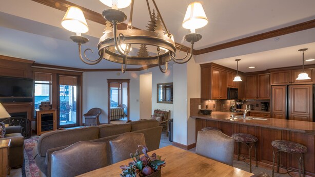 A spacious living area with a wooden dining table, comfortable seating, a chandelier, and a kitchen with wooden cabinets and bar stools.