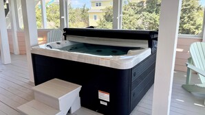 Un bain à remous sur une véranda couverte, accessible par des marches, entouré d’une terrasse en bois et offrant une vue sur des maisons et des arbres.