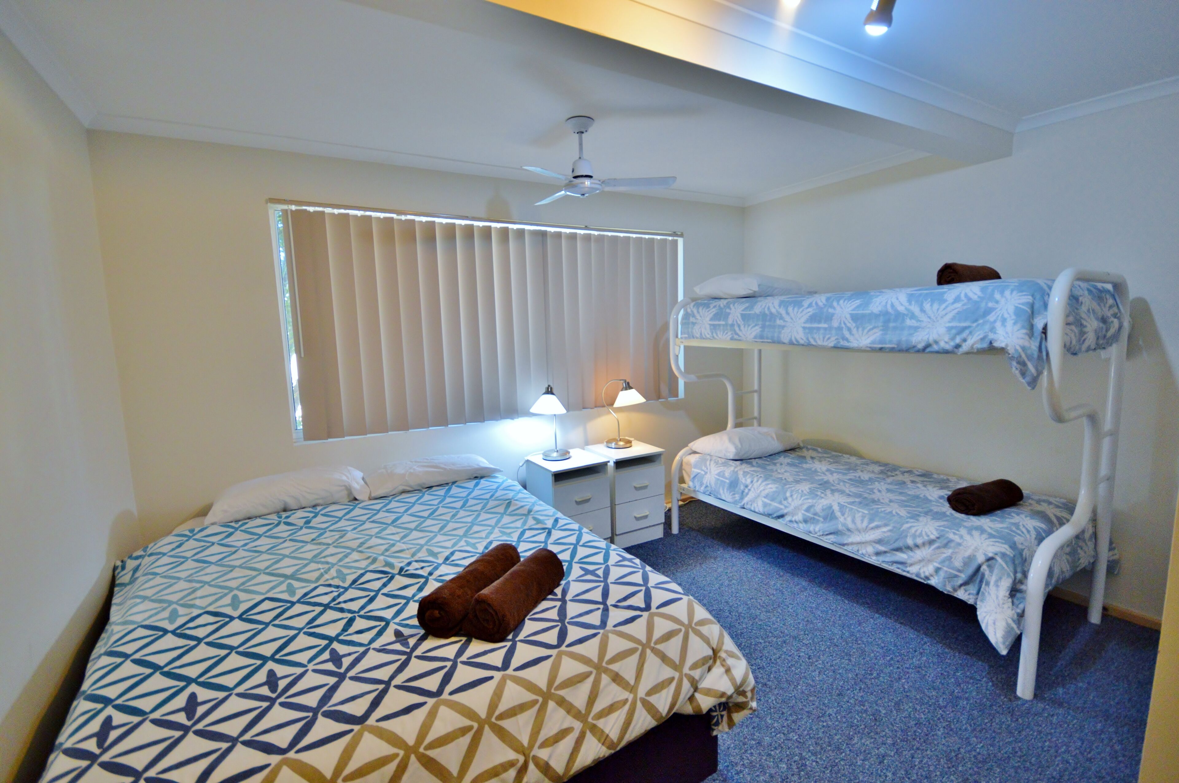 Quarto de hotel com duas camas, um ventilador de teto, uma janela com persianas, mesa de cabeceira com abajur e um tapete azul.