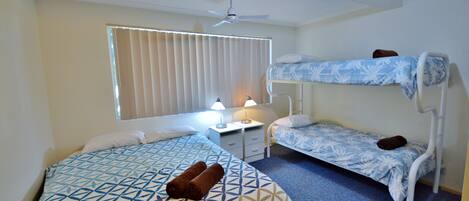 Quarto de hotel com duas camas, um ventilador de teto, uma janela com persianas, mesa de cabeceira com abajur e um tapete azul.