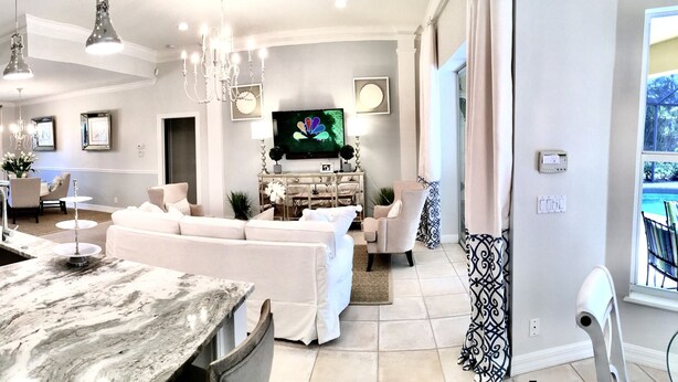 A modern kitchen with white cabinetry, a marble island, and a dining area with a white sofa and a TV. - Exquisite Home in Exclusive Community (Naples)