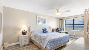 Une chambre à coucher comprenant un lit, deux tables de chevet avec des lampes, un ventilateur de plafond, une fenêtre avec des stores et une chaise.