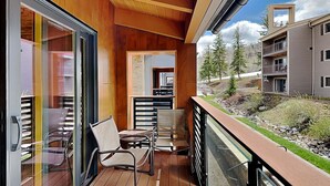 Un balcón con piso de madera, puertas de vidrio y vistas a un edificio moderno y árboles.