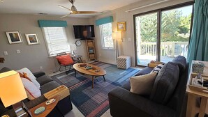 A living room with a sofa, coffee table, and a TV. There are windows with blinds, a ceiling fan, and a view of the outdoors.