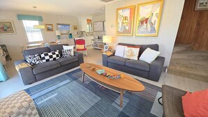 A living room with two grey sofas, a wooden coffee table, and a striped rug. There are framed pictures on the wall and a staircase in the background.