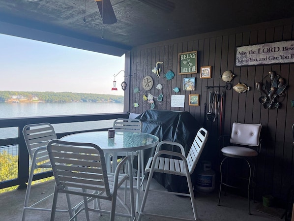 A covered outdoor seating area with a view of a lake, featuring a table, chairs, and various decorative items.