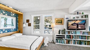 Une chambre à coucher comprenant un lit, une bibliothèque, un téléviseur et une fenêtre donnant sur des immeubles et des arbres.