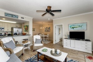 A spacious living room with a large television, wicker furniture, and a dining area in the background.