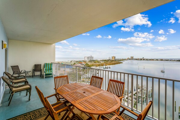A balcony with a wooden table and chairs, overlooking a calm body of water and a cityscape.